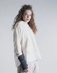 A woman with long curly hair wears the Psophia Cropped Hoodie W/ Shirt Hem and beige cargo pants, styled with dark fingerless gloves, standing against a plain light background.