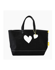 The Tote Imperfect Heart by Kerri Rosenthal is a black cotton canvas bag with two white heart shapes on the front, sturdy black handles, and a yellow side tag, all strikingly contrasted against a plain white background.