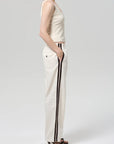 A person stands in profile, hands in pockets, wearing a sleeveless beige top, Citizens Of Humanity/AGOLDE aurora track pant in off white with dark side stripes, and black heeled sandals against a plain light background.