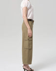 A woman stands in profile against a light gray background wearing Citizens Of Humanity/AGOLDE thassos twisted seam utility pant, a white short-sleeve blouse, dark belt, and black heeled sandals.