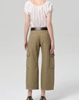 A person stands facing away from the camera, wearing a white short-sleeve blouse and Citizens Of Humanity thassos twisted seam utility pant in khaki, styled with a dark belt and black heeled sandals against a plain light background.