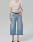Wearing a short-sleeve, light-striped tee, Citizens Of Humanity/AGOLDE vintage culotte in sunlit indigo denim jeans, a brown belt, and black sandals, a person stands against a plain light gray background.