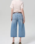 A person stands facing away, wearing Citizens Of Humanity/AGOLDE’s vintage culotte in sunlit with a unique cutout waistband, paired with a striped short-sleeve shirt, black belt, and heeled sandals against a plain light background.
