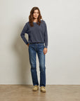 A woman with long brown hair stands against a plain wall, wearing the Perfectwhitetee Ziggy Reverse LS Sweatshirt in dark blue cotton fleece, blue jeans, and beige shoes. She has one hand in her pocket and looks relaxed.