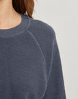 Close-up of a person wearing the Perfectwhitetee Ziggy Reverse LS Sweatshirt in textured dark blue cotton fleece, featuring a crew neckline and bracelet-length sleeves. Only their neck, shoulder, and upper chest are visible against a plain background.