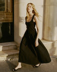 A woman with long blonde hair, wearing DOEN's Sebastiane Skirt as part of a sleeveless black dress and black sandals, walks on a sunlit sidewalk in front of a light-colored stone building.