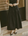 A person wearing the DOEN Sebastiane Skirt— a long black organic cotton skirt with scallop eyelet embroidery— and black open-toe shoes stands on a stone step before a building with a black door and pillar. Their upper face is not shown.