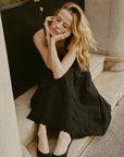 A woman with long blond hair in a black dress and DOEN's Sebastiane Skirt sits peacefully on a stone step, resting her chin on her hand with eyes closed.