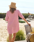 A woman in the Frank & Eileen MARY Shirtdress S25 and a straw hat stands on the beach, holding a wooden chair with a woven bag and towel—capturing effortless seaside style.