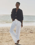 A woman stands barefoot on the beach, smiling, wearing a loose black sweater with large buttons and Frank & Eileen’s Limerick Carpenter Pant. The ocean and sky form the background.