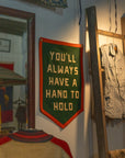 The Oxford Pennant "You'll Always Have a Hand to Hold" camp flag in green and orange adds a cozy touch to your space, displayed near a wooden ladder, striped shirt, and mirror in a warmly lit room.