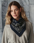 A woman with wavy, light brown hair wears a grey top and The Bandana Kerchief by The Great Inc.—a dark blue alpaca accessory with a white paisley design—around her neck as she stands in front of a textured grey fabric background.
