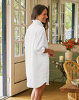 A woman wearing the Frank & Eileen RUFFLE MARY Classic Shirt Dress F25 smiles indoors by an open door. The room features flowers, a lit candle, and rattan chairs, with greenery visible outside.