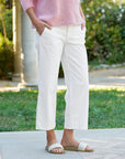 A person wearing a pink sweater and Frank & Eileen's Westport Italian wide-leg chinos is outdoors on a stone path, hands in pockets, with beige slide sandals. The backdrop is a garden featuring green grass and a stone pillar.