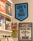 A record store wall features Oxford Pennant’s bright blue “Life’s So Rad” flag, shelves of vinyl, a vibrant cartoon poster, and a black-and-white sign for vintage vinyl and CDs upstairs—ideal dorm room decor inspo.
