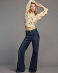 A person poses with arms raised, wearing FRAME’s The Uniform Trench—a lightweight, tailored beige cropped jacket—over high-waisted dark blue wide-leg jeans, set against a plain grey background.