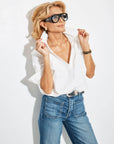 A stylish blonde woman in large black sunglasses and blue jeans poses against a white background, confidently adjusting the collar of her 100% cotton Classic Collared Henley, the Celeste Parachute by A Shirt Thing.
