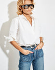 A woman with blonde hair and large black glasses wears the Celeste Parachute white button-up shirt by A Shirt Thing and blue jeans, with her hands in her pockets, standing against a plain white background.