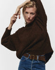 A person models the Janessa Leone Bowe Belt, styled with a textured dark brown sweater and blue jeans, striking a confident pose against a plain white background.