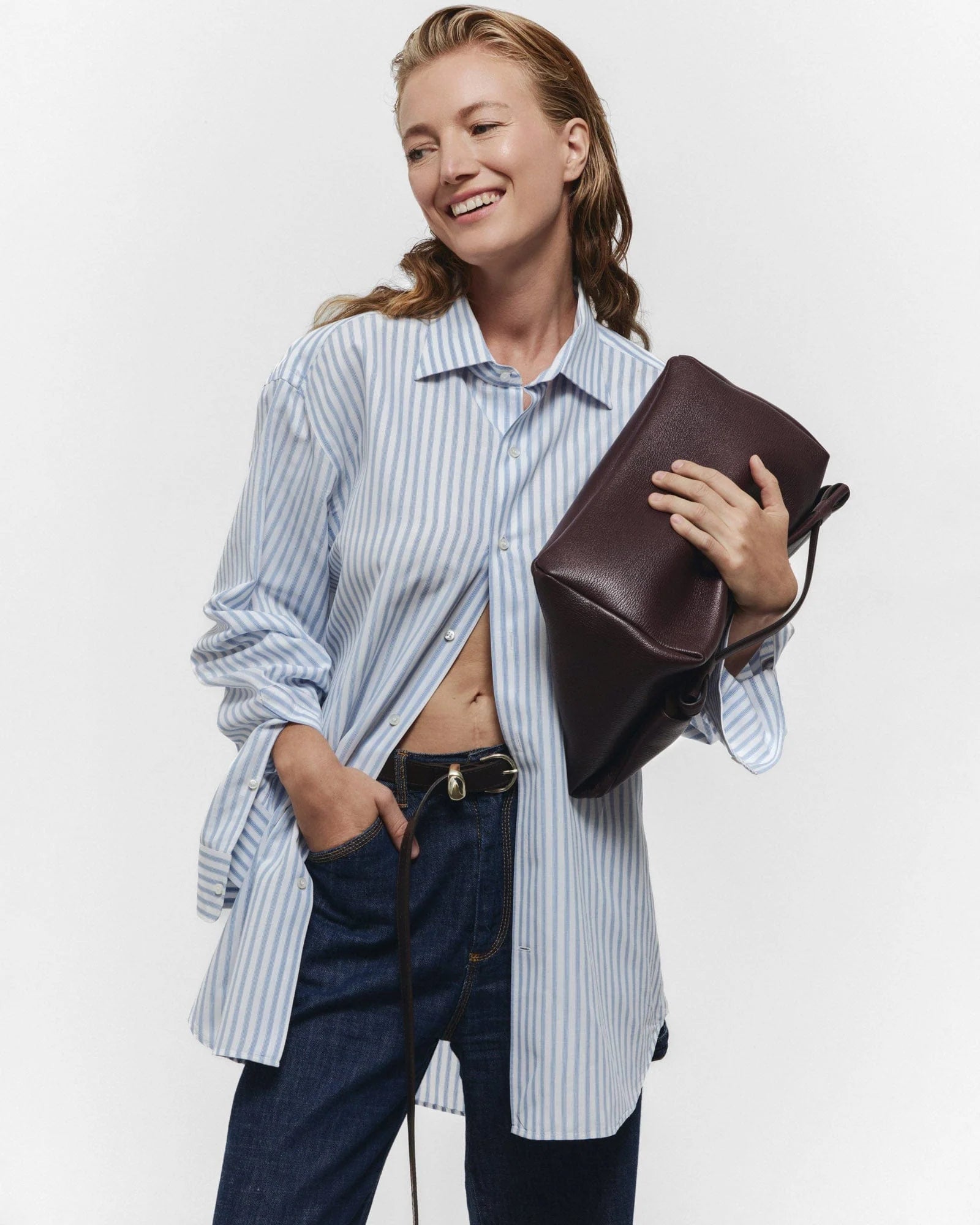 A woman in a blue and white striped button-up shirt and dark jeans smiles while holding the Janessa Leone Myra DW Belt under her arm against a plain white background.