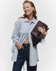 A woman in a blue and white striped button-up shirt and dark jeans smiles while holding the Janessa Leone Myra DW Belt under her arm against a plain white background.