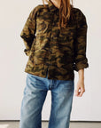 Model wearing The Work Shirt by Hey Gang in a boxy fit camouflage design with rolled-up sleeves, paired with blue jeans. Photographed against a plain light background; head is partially out of frame.