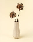A white, textured ceramic vase holds two Indaba Dahlia Stem Russet faux flowers with long, slender stalks, serving as minimal accent decor against a plain, light beige background.