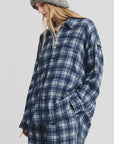 A woman with long blonde hair wears the Denimist BUTTON FRONT SHIRT in blue/ecru plaid, styled with matching pants and a chunky gray knit beanie. She stands against a white background with one hand in her pocket.