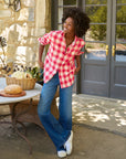 A woman in the Frank & Eileen Eileen Button Up Shirt F25, styled in red and white plaid, pairs it with blue jeans and smiles outdoors beside a table of bread and vegetables, set before double glass doors and a stone wall.