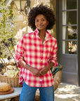 A woman stands outside by a stone wall and glass door, smiling gently in the Frank & Eileen Eileen Button Up Shirt F25, a red and white plaid style, paired with blue jeans. A basket of bread and vegetables sits on a nearby table.