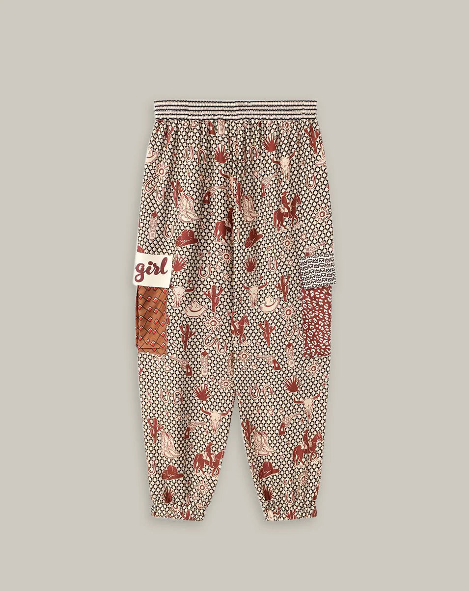 The Gianna Cargo Pants by Me369 are loose-fitting with an elastic waist, an abstract red, beige, and black rodeo print, a white "girl" patch, and a red checkered patch on the side, shown against a plain background.
