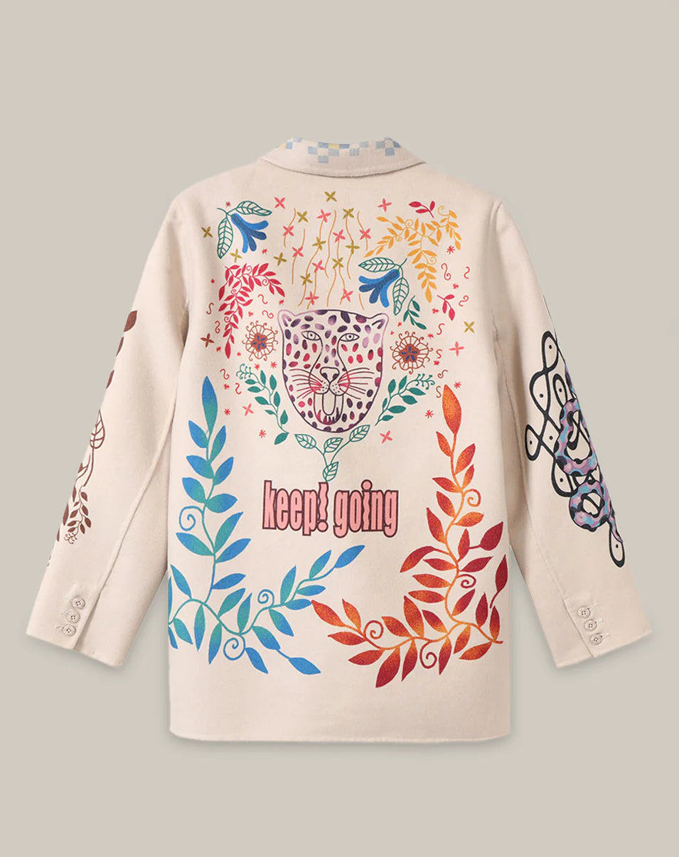 The Juliana Wool Jacket by Me369 is an off-white, long-sleeve blazer with colorful, whimsical designs—leopard face, florals, swirling shapes, and “keep! going” on the back—featuring red, blue, yellow, and purple accents.