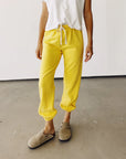 A person in a white t-shirt and yellow Hey Gang "The Sweatpants" joggers with an elastic waistband stands on a concrete floor. They pair beige suede clogs against a plain white wall.