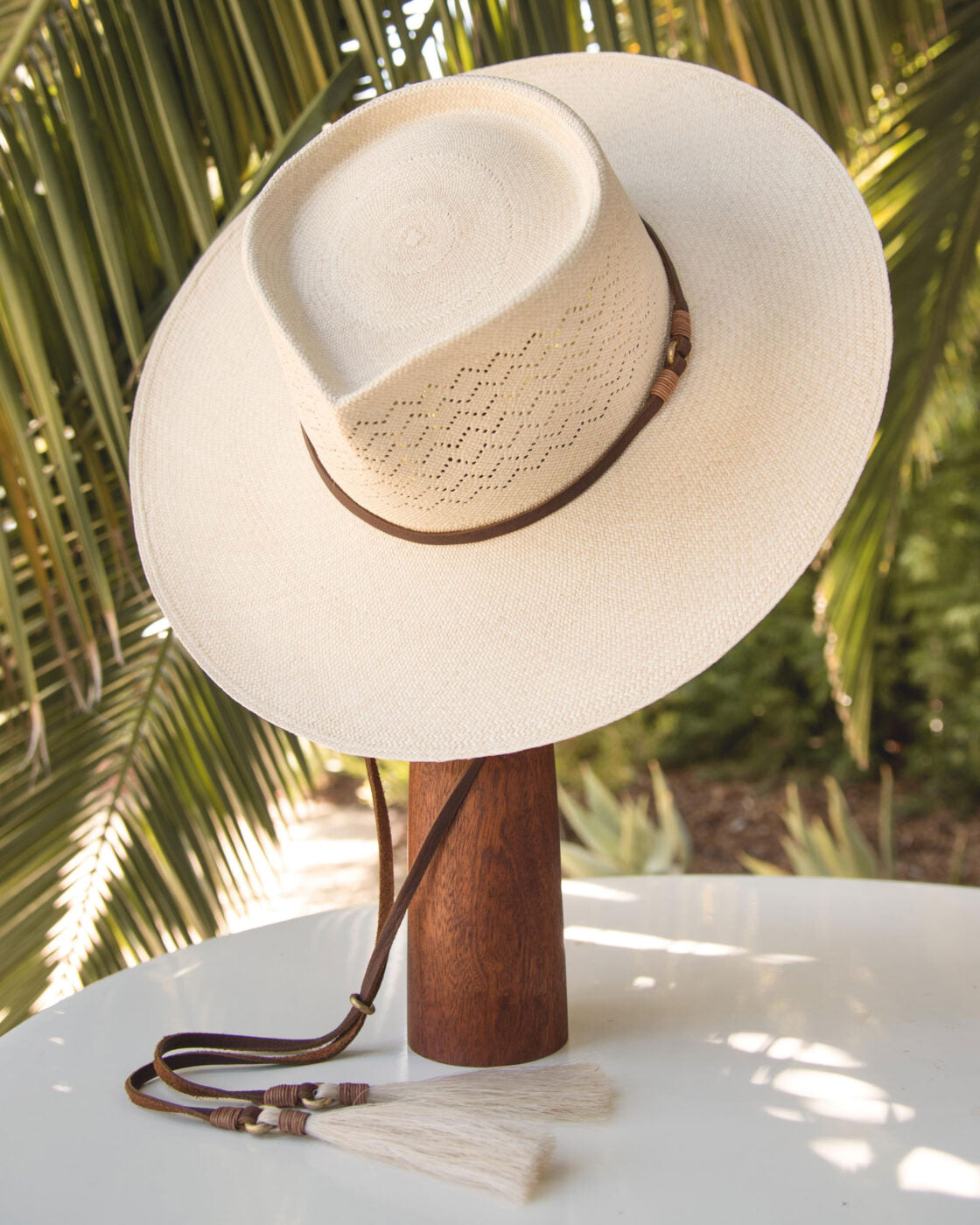 A wide-brimmed white Ninakuru Toquilla straw Paloma Hat with a brown leather band on a wooden stand, against a background of green palm fronds.