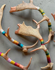 The Faire Wool & Copper Wrap Deer Antler Fallow features handcrafted antlers on a gray surface, each partly wrapped in colorful yarn, creating vibrant, unique wall decor.