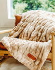 A plush Cascade Throw Blanket by Faire, ruched for extra texture, is draped over a wooden chair with tan leather cushions near a window overlooking greenery. A leather tag on the blanket displays the brand name "Faire.