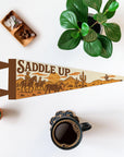 The "Saddle Up" Felt Pennant by Faire, featuring a cowboy scene, rests on a white surface with coffee, green plants, and crystals—ideal as western decor or a gift for horse lovers.