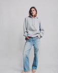 A person stands against a light background wearing a loose grey hoodie, Denimist LUCY BF JEAN MAIRE BLUE boyfriend jeans, and white sandals, hands in pockets and looking relaxed.