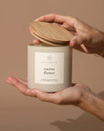 A person holds the Faire "Cactus Flower - 14oz. Candle" in one hand and lifts its wooden lid with the other, against a plain light brown background.