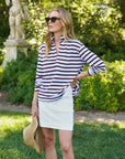 A woman outdoors holds a straw hat, wearing sunglasses, a white skirt, and the Frank & Eileen Patrick Popover Henley. Greenery and a statue are visible in the background.