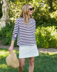 Wearing the Frank & Eileen Patrick Popover Henley, a woman in sunglasses and a white skirt stands on grass, holding a floppy straw hat. She poses in front of green bushes and a large statue under the sun.