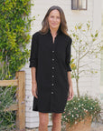 A woman with long brown hair stands outside, smiling in the Frank & Eileen Mary Classic Shirtdress–Ruffle, featuring rolled-up sleeves and a flattering fit. Greenery and a potted plant provide a fresh, natural backdrop.
