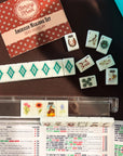 Faire Wild West Mahjong SET, featuring tiles (with jokers and character tiles), 4 racks, a bag, and a mat displayed on a brown surface with the set box, rules card, and arranged rows of tiles visible.