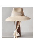 The Tatum Extra Long Brim Hat by Ninakuru is made from toquilla straw and rests on a wooden stand. It boasts a beige design with a textured fabric band around the crown and long ties knotted at the back, all set against a smooth, light background that elevates its sustainable elegance.