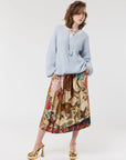 A woman stands against a plain white background wearing a loose light blue sweater, the Me369 Vanessa Midi Skirt with jungle animal prints, gold high-heeled sandals, and layered necklaces.
