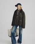 A person stands against a plain background wearing Denimist's O/S Field Jacket, a navy cap, striped shirt, flared jeans, sneakers, and holding a large white tote bag.