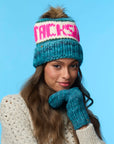 A woman wearing the Faire Knit Ski Hat—blue merino wool with faux fur pom pom and bold pink lettering—plus matching mittens and a cream sweater, poses against a blue background.