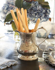 The Vintage Pitcher with Deer by Faire, crafted in Italy, holds breadsticks on a set dining table. Blue hydrangeas add a decorative touch in the background, alongside ruffled napkins and neatly stacked plates.