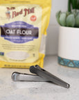 A Faire Stainless Steel Kitchen Bag Clip sits on a countertop in front of a yellow bag of Bob's Red Mill oat flour, with a potted succulent plant in the background.