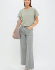 A woman with straight brown hair and a slight smile stands against a plain background, wearing the ASKK Cuff Tee in light green (100% cotton), light gray wide-leg jeans, beige sneakers, and a taupe shoulder handbag. Made in Los Angeles.